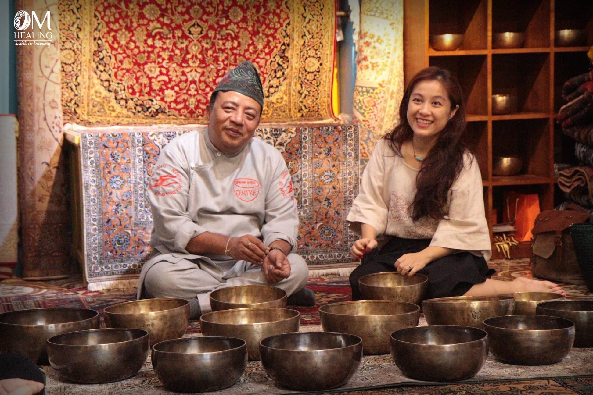 Master Santa Ratna Shakya và Founder Hiền Trần ngồi bên hàng chuông xoay, nền thảm Tây Tạng đỏ.